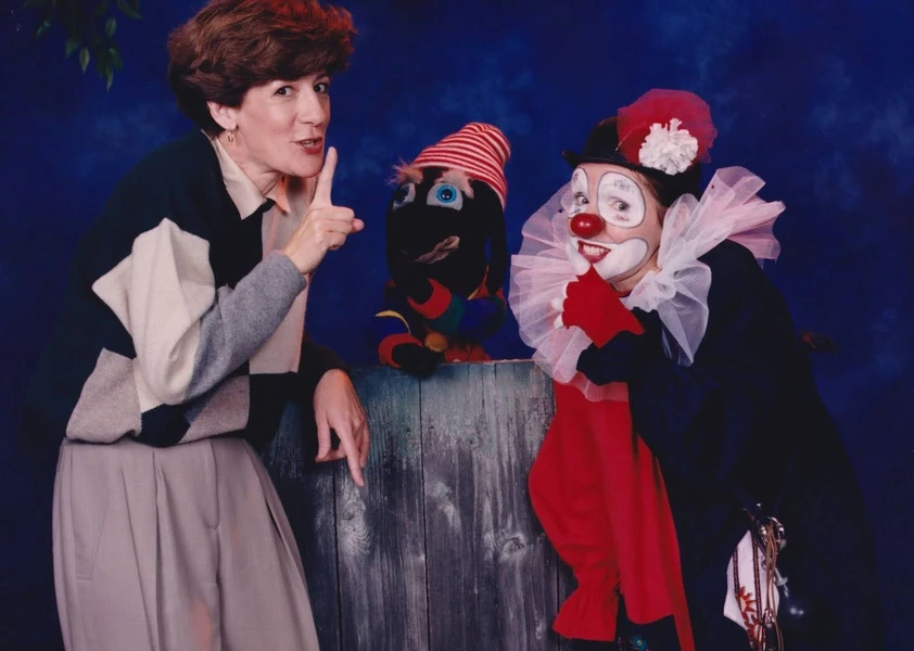 Une femme fait un geste de silence avec le doigt sur les lèvres, un personnage marionnette coloré apparaît au centre, et un clown maquillé en rouge, blanc et noir sourit en imitant le même geste.